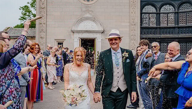 Bride and groom leaving Bourton Hall wedding ceremony surrounded by guests throwing confetti, wearing Fraser Hart wedding rings